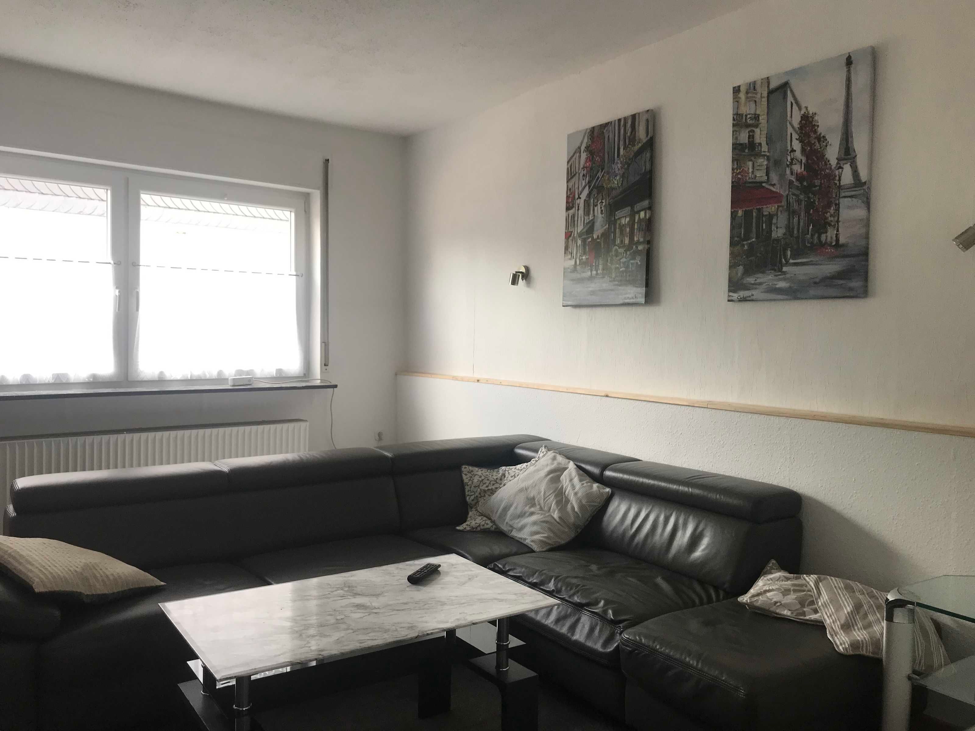 Un salon moderne avec un canapé en cuir, une table basse en verre et deux tableaux représentant des paysages urbains, encadrés et accrochés au mur.