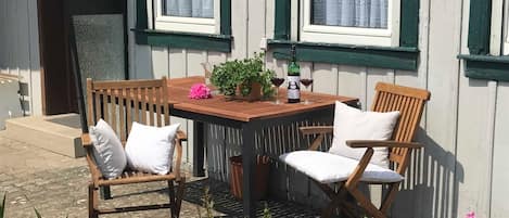 A wooden table with two chairs, a potted plant, and a bottle of wine on a balcony with white walls and green window frames.