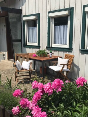 A wooden table with two chairs, a potted plant, and a bottle of wine on a balcony with white walls and green window frames.