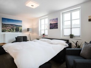 Un dormitorio con una cama grande, dos ventanas, un radiador, una lámpara de pie y un cuadro en la pared.