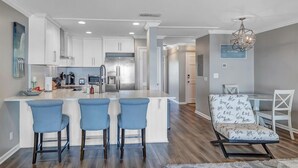 A modern kitchen with white cabinets, a central island with blue bar stools, and a dining area with a patterned chair. - Wellness Retreat - Oceanfront condo w beach access (Ponte Vedra Beach)