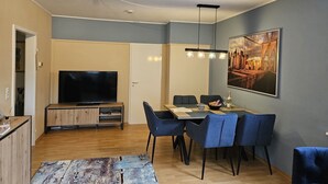 Un salon moderne avec un téléviseur à écran plat, des meubles en bois, une table à manger avec des chaises bleues et un grand tableau représentant un paysage urbain accroché au mur.