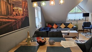 Un salon moderne avec un tableau représentant un paysage urbain, un canapé agrémenté de coussins colorés, une table à manger avec une bouteille et des verres, et un luminaire suspendu au plafond.
