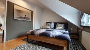 Une chambre avec un plafond en pente, un lit avec une literie à motifs, un grand tableau et une fenêtre avec des rideaux.