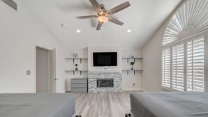 A modern bedroom with a ceiling fan, a fireplace, a TV mounted above it, and a large window with blinds. - Cozy 5-Bedroom Oasis Near the Strip: Arcades, Pool, and BBQ Haven! (Las Vegas)