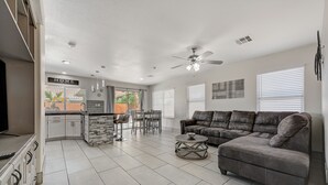 A spacious living area with a gray sofa, a dining table with chairs, a kitchen area, and a ceiling fan. - Cozy 5-Bedroom Oasis Near the Strip: Arcades, Pool, and BBQ Haven! (Las Vegas)