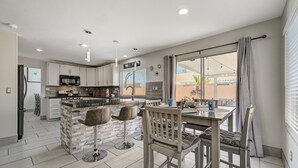 A modern kitchen with a central island, bar stools, and a dining table with chairs. - Cozy 5-Bedroom Oasis Near the Strip: Arcades, Pool, and BBQ Haven! (Las Vegas)