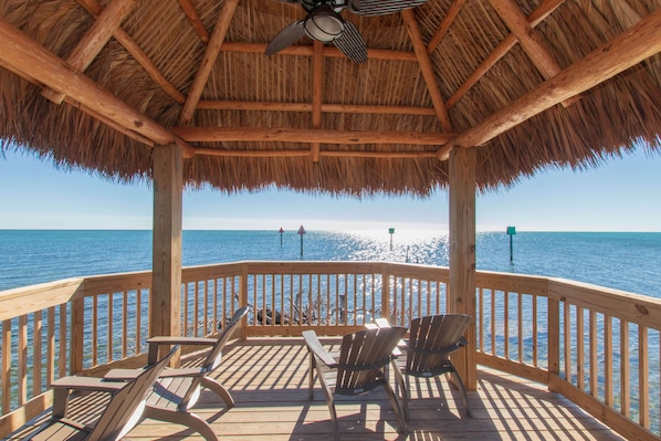 A wooden deck with chairs and a thatched roof, overlooking the sea.