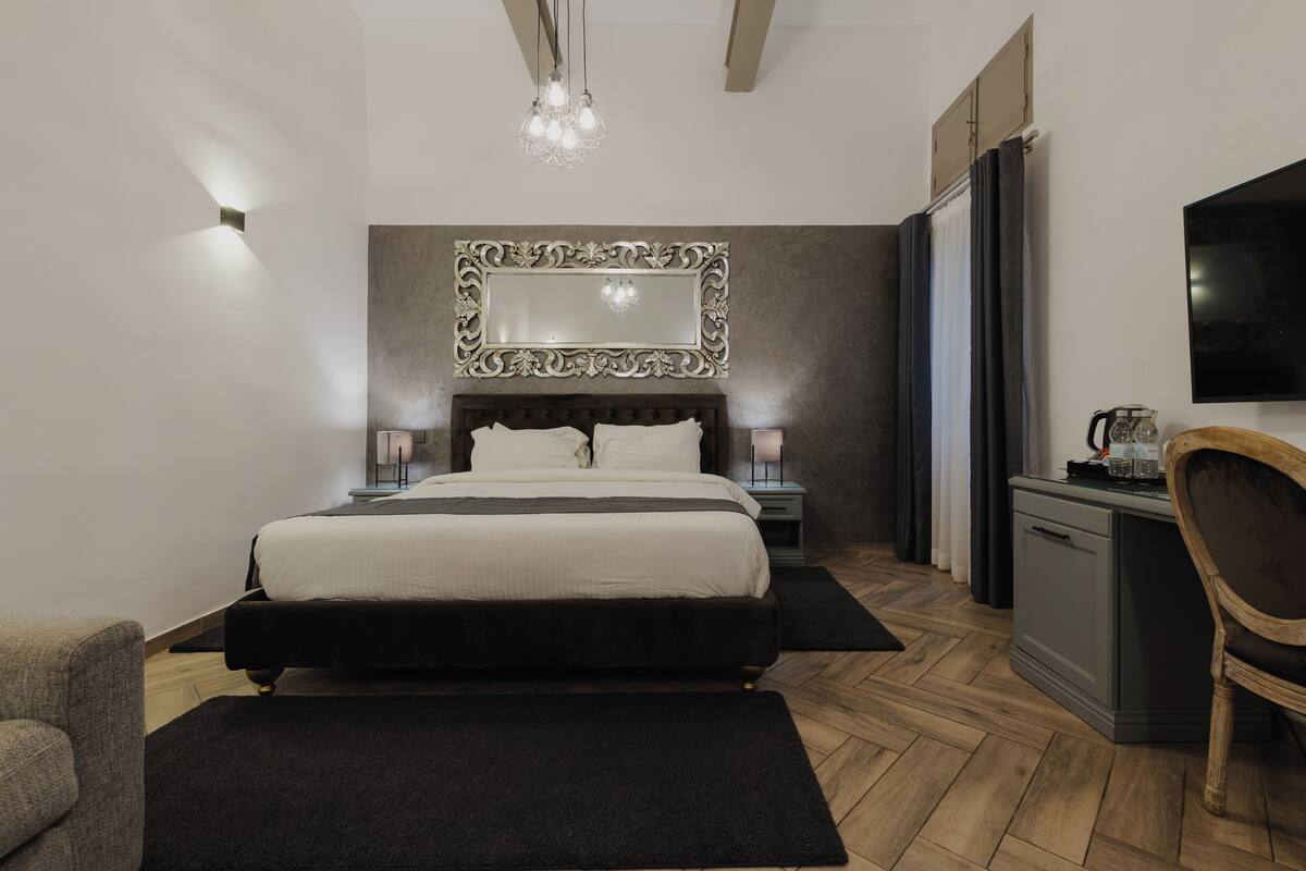 A modern bedroom with a large bed, a mirror above it, bedside lamps, a desk with a chair, and a television on the wall.