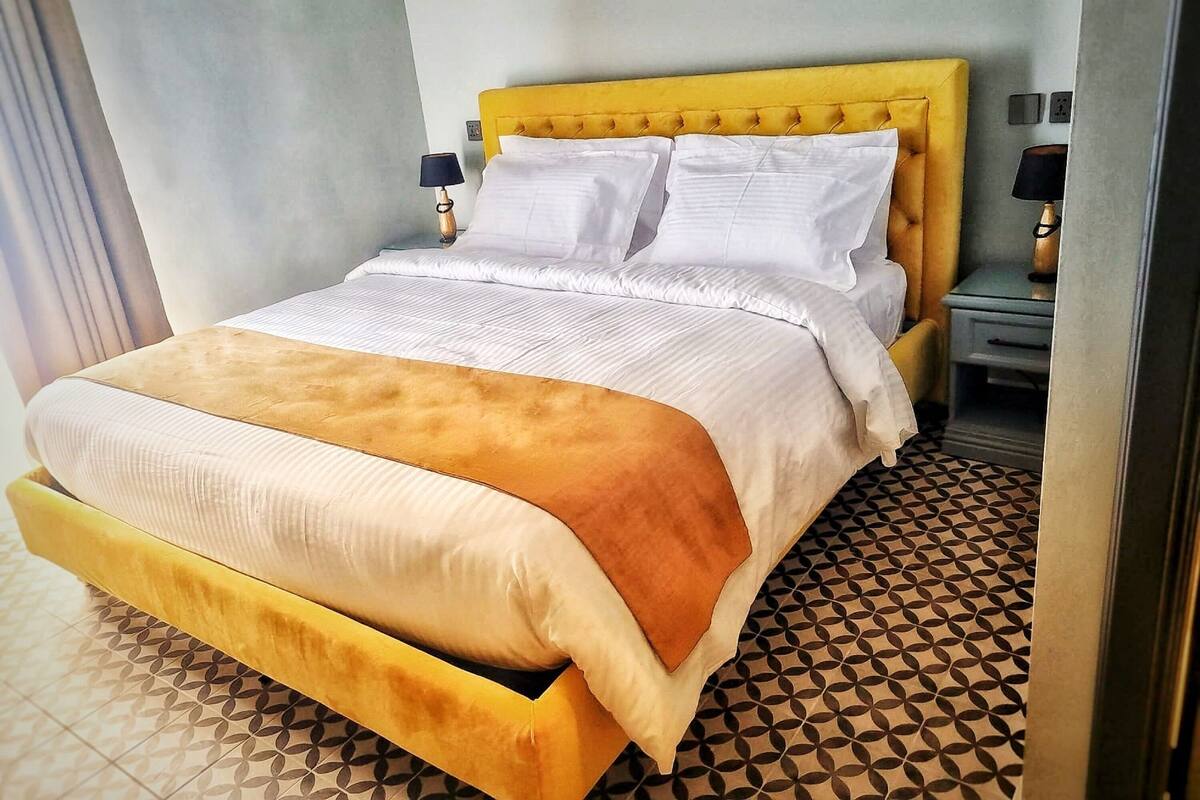 A bed with a yellow headboard, white bedding, and a brown throw blanket. There are two bedside tables with lamps. The floor has a geometric patterned tile design.