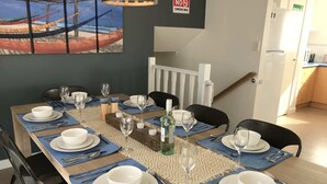 A dining table set for six with blue placemats, white plates, and silverware. A painting of a beach scene hangs on the wall above the table. - Bay Views Beach Surfing Fishing Boating (Surf Beach)