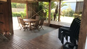 A covered outdoor patio with a wooden table and chairs, surrounded by plants and a view of the outdoors. - Bay Views Beach Surfing Fishing Boating (Surf Beach)