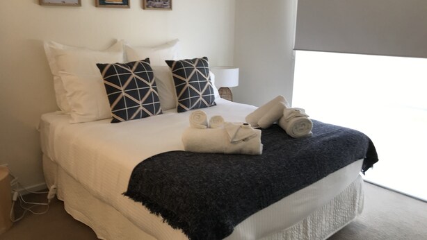 A neatly made bed with a dark blue throw blanket, white pillows, and a rolled towel on the side. - Bay Views Beach Surfing Fishing Boating (Surf Beach)