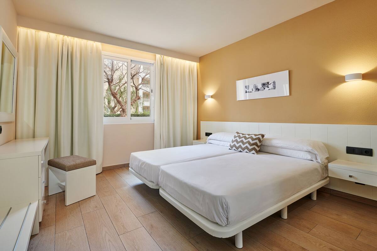 Una cama doble con cabecera, mesitas de noche, un taburete pequeño y una ventana con cortinas.