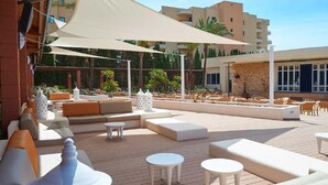 An outdoor lounge area with multiple seating options including sofas and tables, shaded by large umbrellas, and surrounded by greenery and buildings. - Protur Badí­a Park Aparthotel (Sant Llorenc des Cardassar)