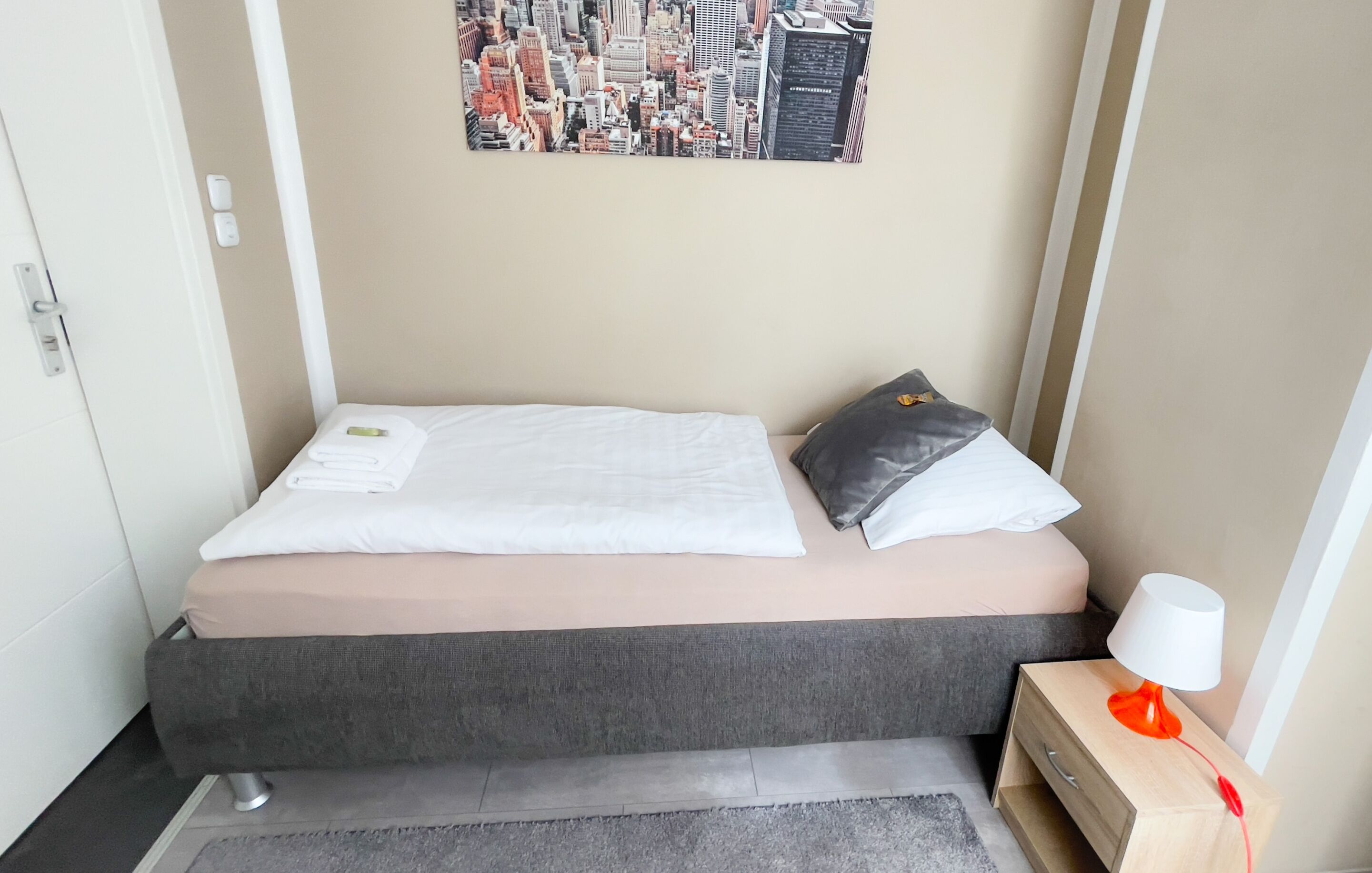 A small, neatly arranged bedroom with a bed, a nightstand, a lamp, and a cityscape poster on the wall.
