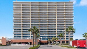 A multi-story hotel with a uniform exterior, featuring a central entrance with a covered walkway, palm trees, and a red truck parked nearby.