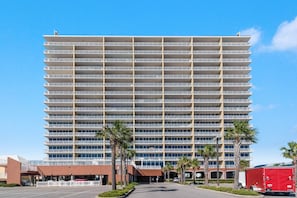 A multi-story hotel with a uniform exterior, featuring a central entrance with a covered walkway, palm trees, and a red truck parked nearby. - Sanibel 402 | Gulf View | Outdoor Pool, Hot Tub, & Sauna | My Beach Getaways (Gulf Shores)