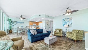 A spacious living room with a blue sofa, green armchairs, and a glass coffee table. There is a dining area with a table and chairs, and a kitchen in the background.