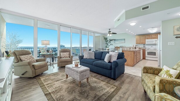 A spacious living room with a blue sofa, beige armchairs, and a patterned rug. A kitchen with wooden cabinets and a microwave is visible in the background.