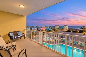 A balcony with a pool, a wicker sofa, and a view of a coastal town at sunset.