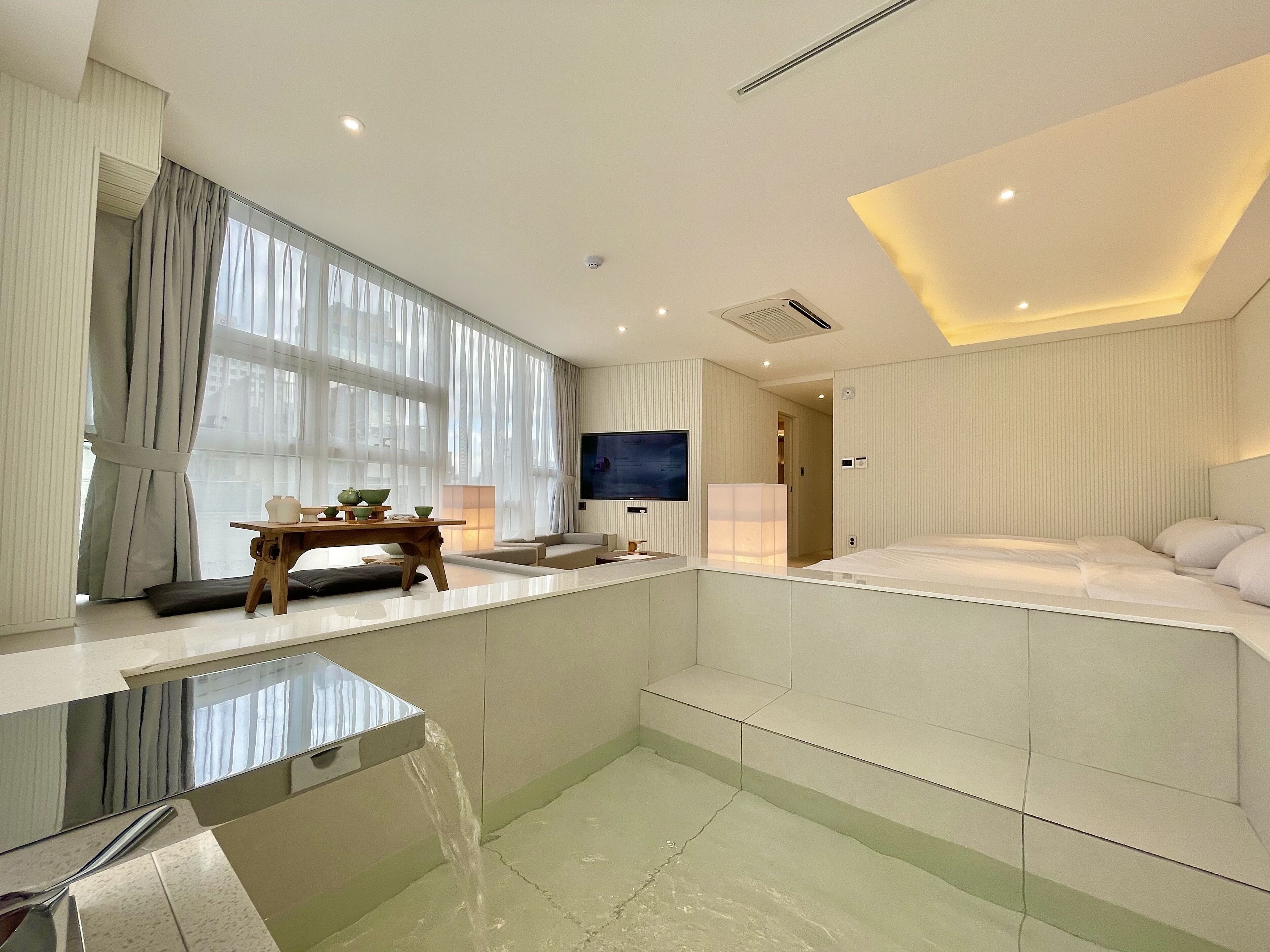 A modern hotel room with a large bed, a TV, a small table with a bowl, and a bathroom with a sink and mirror.