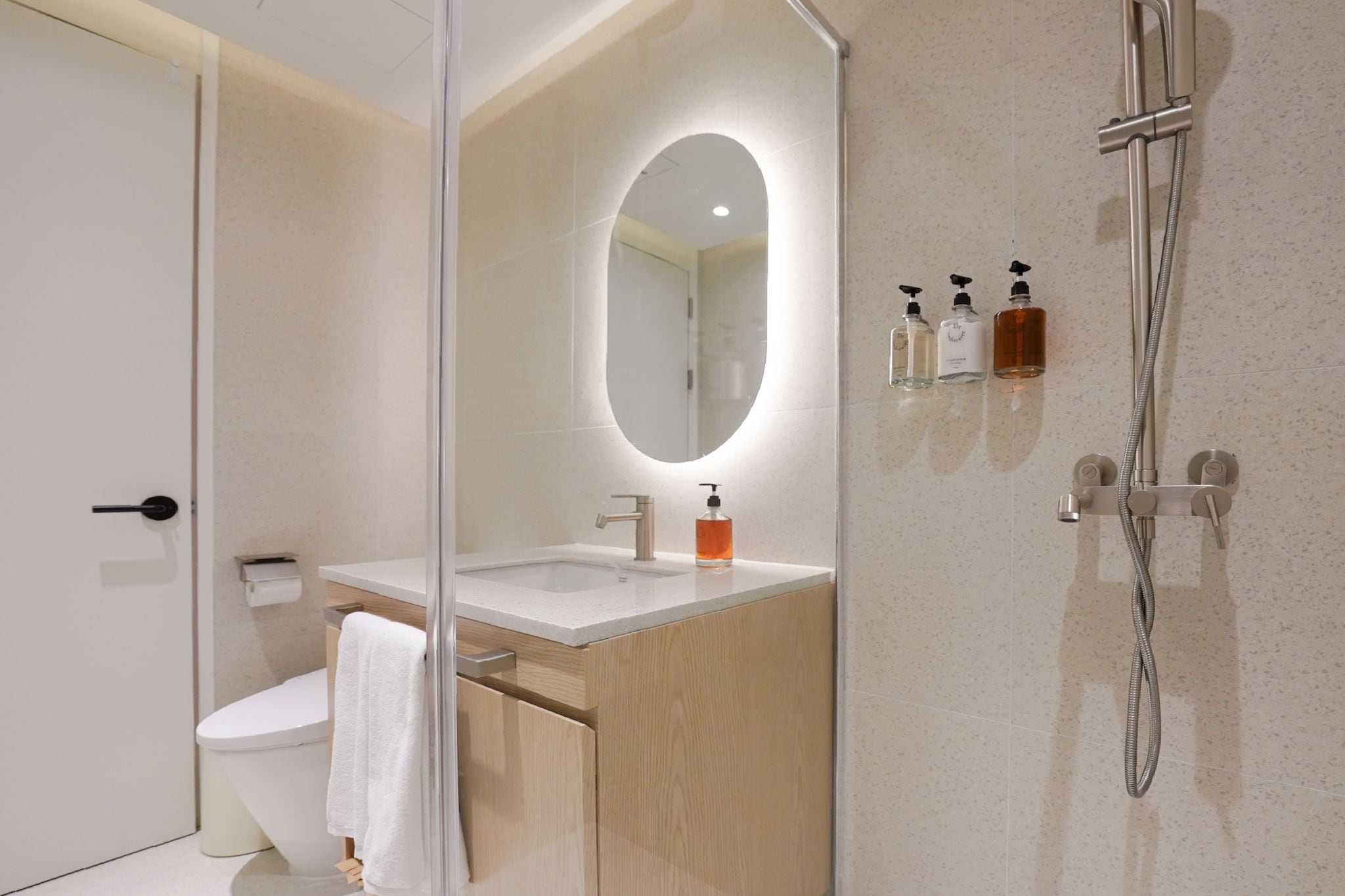 A modern bathroom with a glass shower enclosure, a white toilet, a sink with a mirror, and three bottles of soap on the wall.