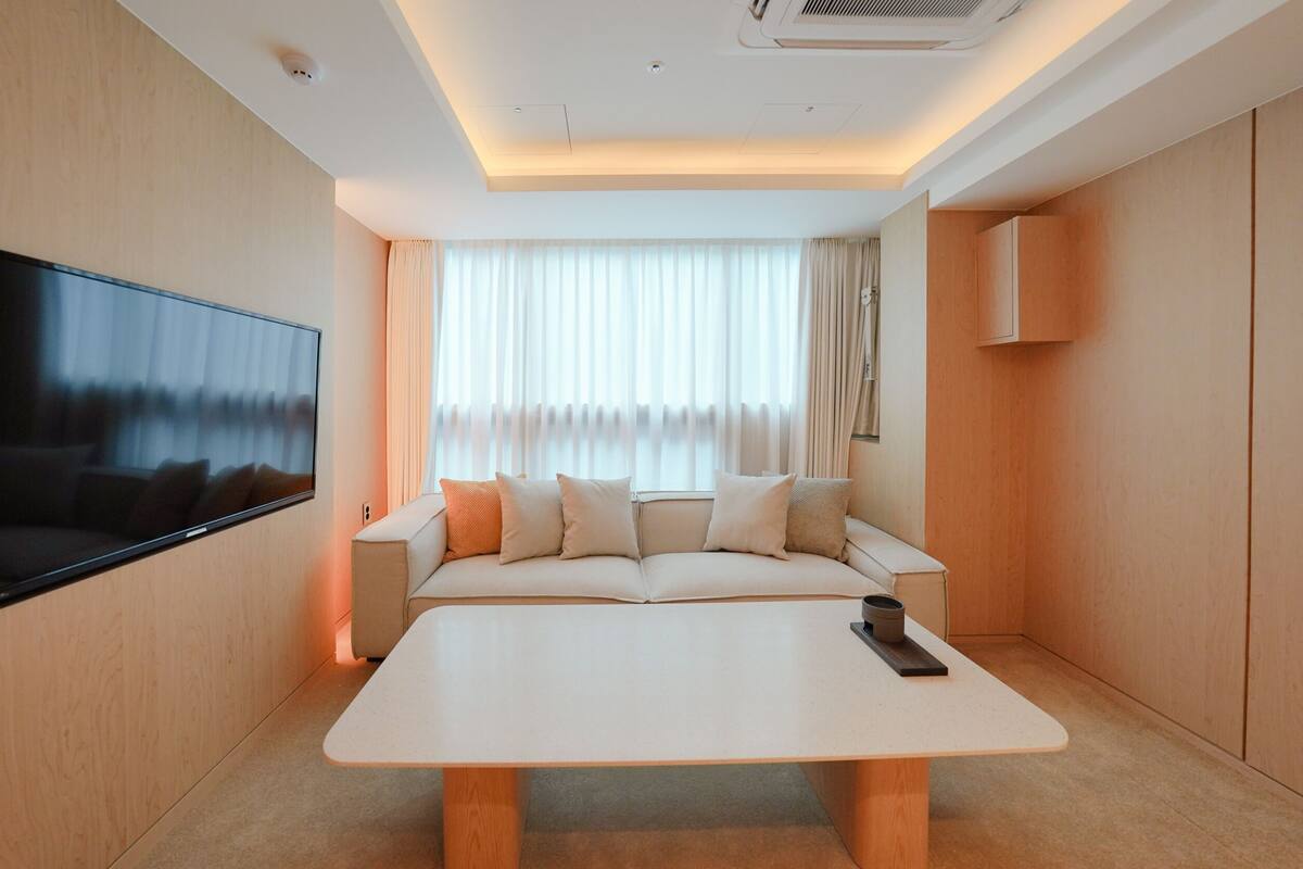 A modern hotel room with a large flat-screen TV mounted on the wall, a sofa with cushions, a coffee table, and a window with curtains.