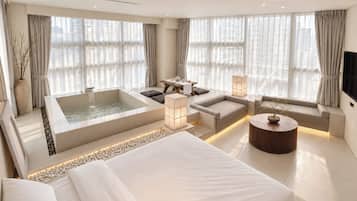A modern hotel room with a large bed, a sitting area with a table and chairs, a bathtub, and large windows with curtains.