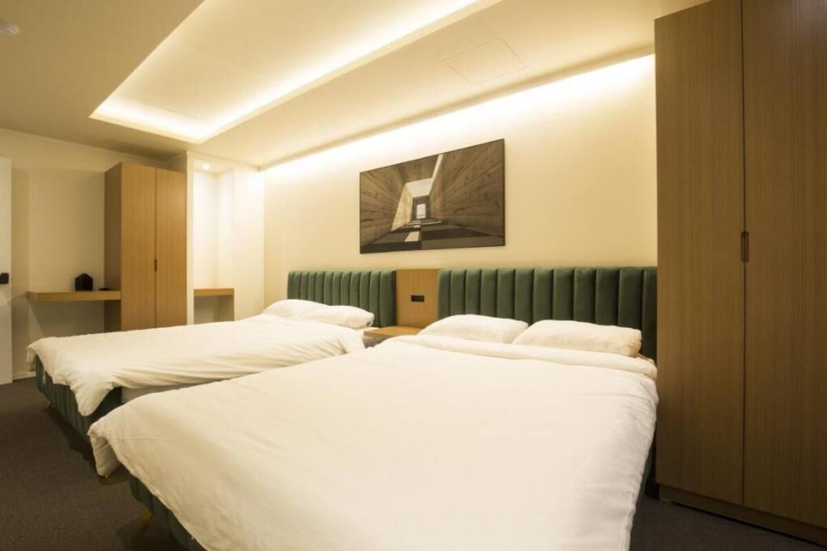 A hotel room with two beds, a green upholstered headboard, a wooden wardrobe, and a framed artwork on the wall.