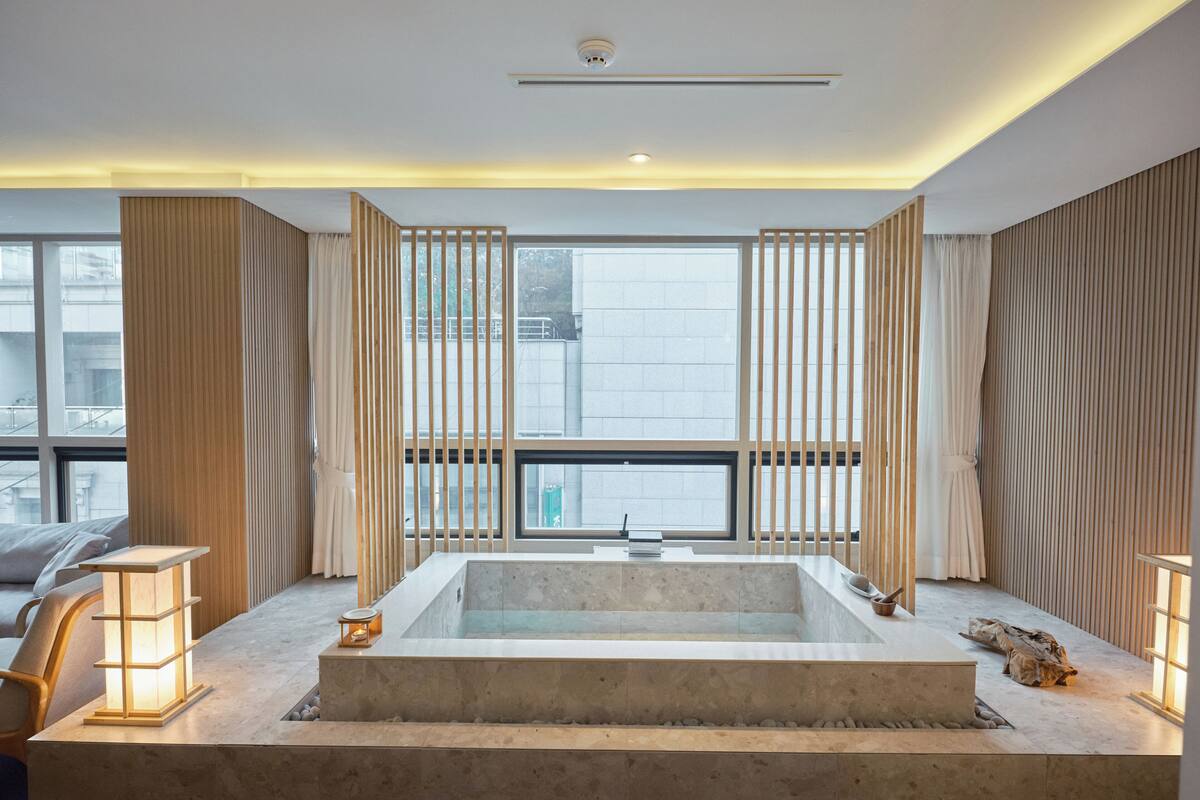 A modern bathroom with a large bathtub, wooden panel dividers, and a view of the city through large windows.