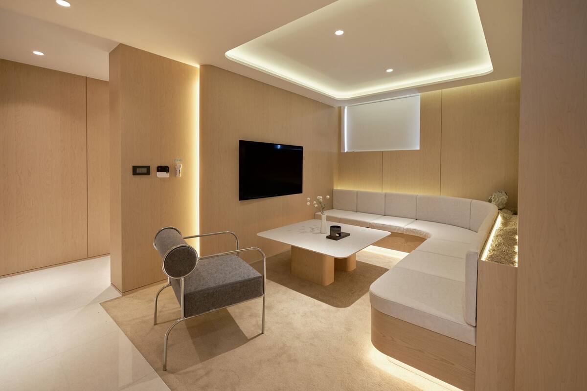 A modern, well-lit room with a flat-screen TV mounted on the wall, a comfortable seating area with a sofa and a cushioned bench, and a minimalist chair with a metal frame.
