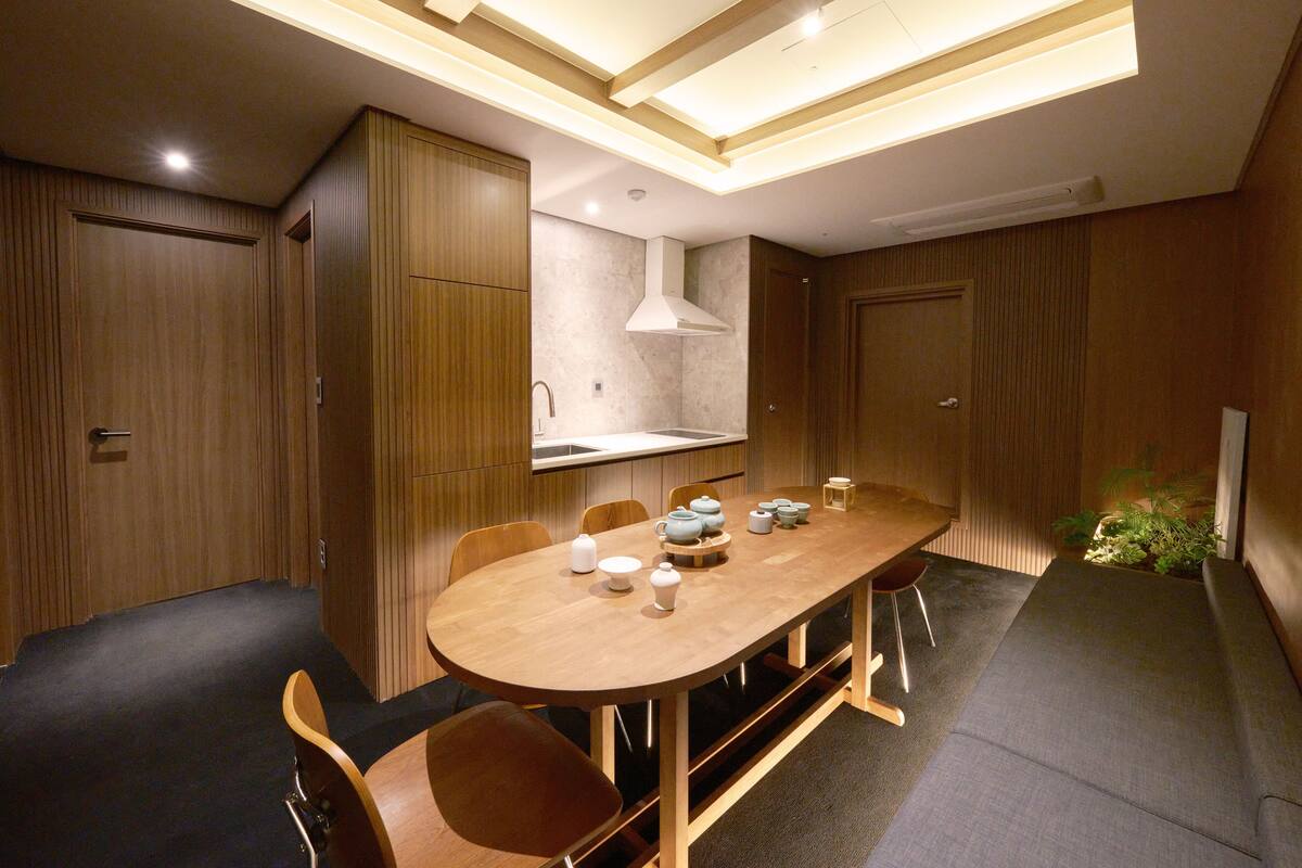 A modern dining area with a wooden table and chairs, a kitchen in the background, and a small plant on the right.
