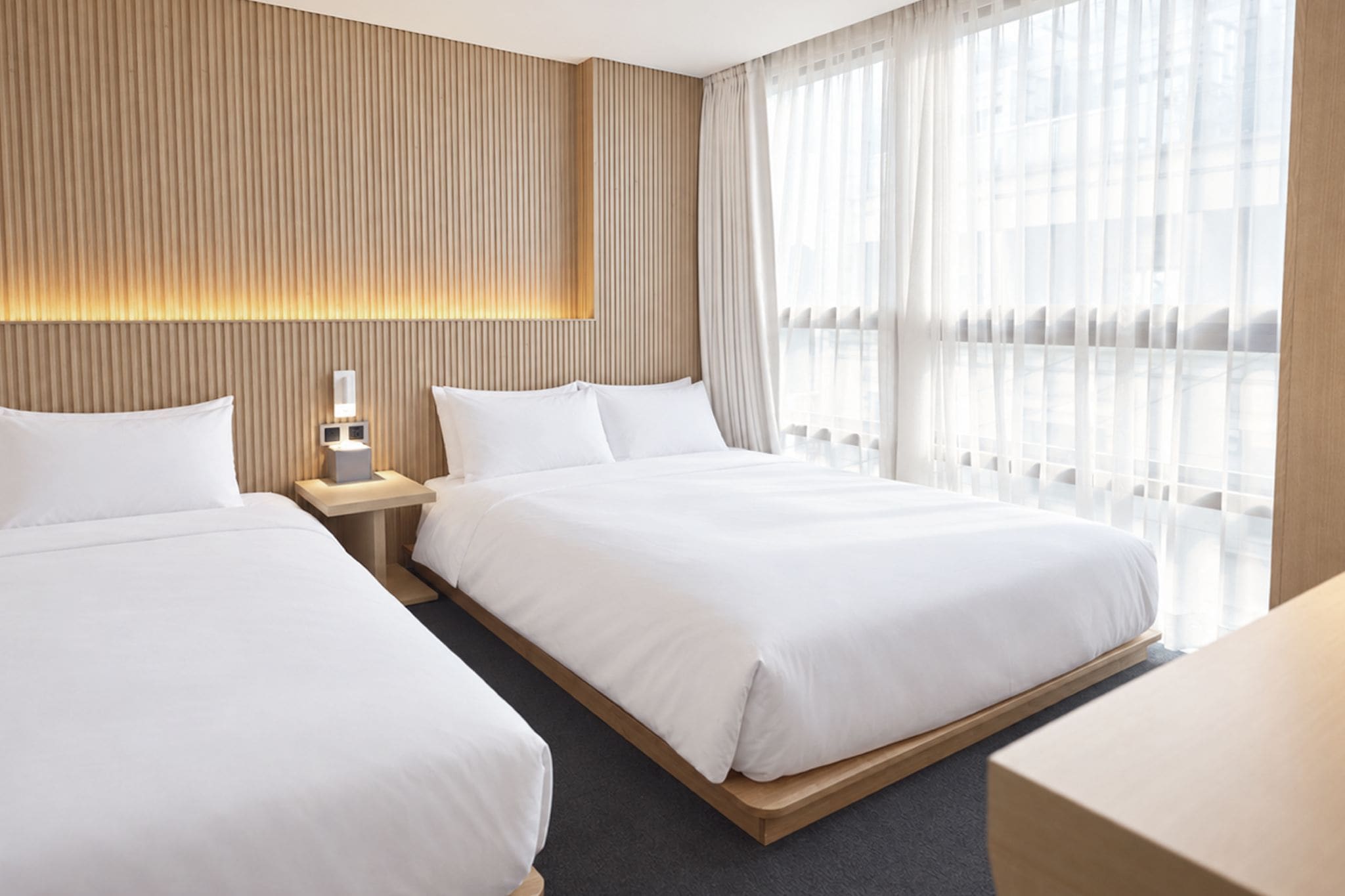 Quarto de hotel com duas camas, mesa de cabeceira e uma janela grande com cortinas.