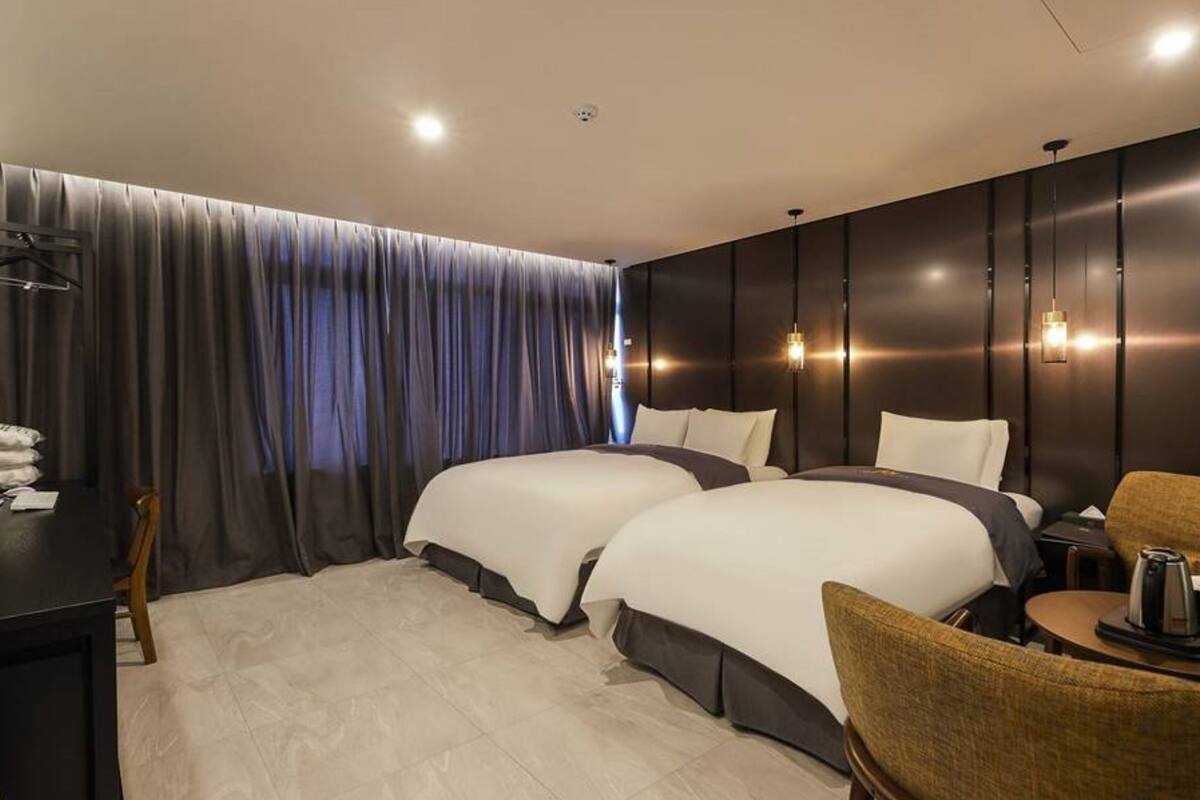 A hotel room with two beds, a desk, a chair, and a small table with a kettle.