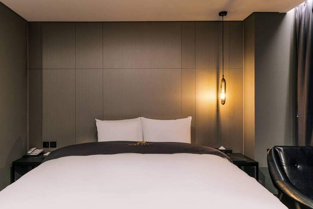 A modern hotel room with a large bed, a bedside table, a chair, and a wall-mounted light fixture.