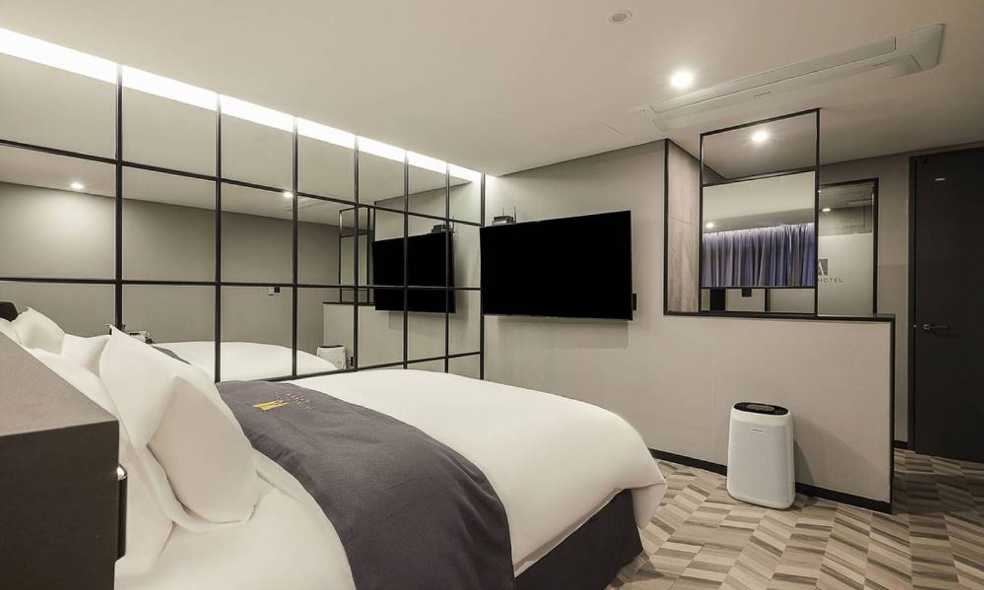 A modern hotel room with a large bed, a flat-screen TV mounted on the wall, and a mirror reflecting the room.