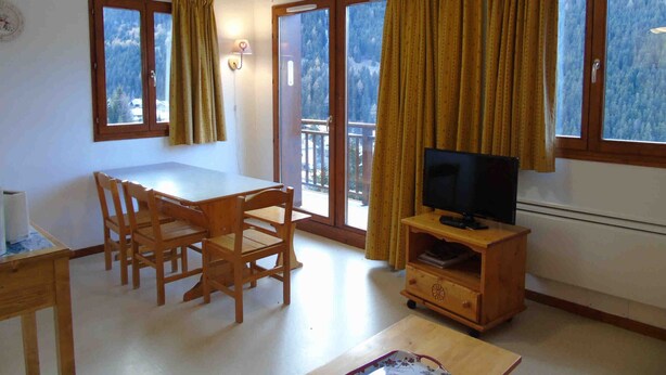 A room with a dining table and chairs, a TV on a wooden stand, and large windows with curtains. - Cozy apartment in Valfréjus, near lifts (Modane)