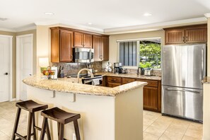 A modern kitchen with wooden cabinets, a stainless steel refrigerator, a granite countertop, and a window overlooking a garden. - 🏖️Mauna Lani Golf Villas H22 | Golf Course Views + Private Beach Club (Kamuela)