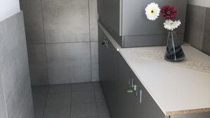 Ein Badezimmer mit grauen Fliesen, einem weißen Waschbecken und einem Schrank. An der Wand befindet sich eine kleine Lüftung und ein Abfluss im Boden.