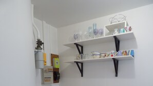 Une petite cuisine avec une table en bois et deux chaises, une étagère où sont rangés des verres, et un sol carrelé.