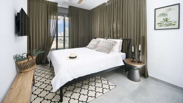 Un dormitorio moderno con cama, ventilador de techo, televisor montado en la pared, mesita de noche con velas y ventana con cortinas.