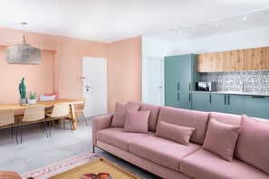 A modern living room with a pink sofa, wooden dining table, and a kitchen with teal cabinets.