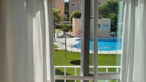 A view through a window with white curtains, revealing a swimming pool, a grassy area, and buildings in the background. - IN THE QUIET AREA OF NOVO SANCTI PETRI (Chiclana de la Frontera)
