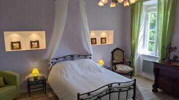 A bedroom with a four-poster bed, a green armchair, a wooden dresser, and a window with green curtains.