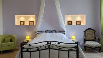 A bed with a canopy, two bedside lamps, a green armchair, a floral-patterned chair, and wall-mounted picture frames.