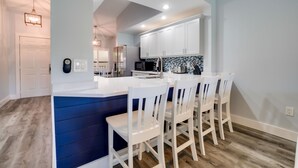 A modern kitchen with white cabinets, a blue accent wall, and a central island with white bar stools. - Near IMG - Shorewalk Unit 202 by SeaBreeze Vacation (Bradenton)