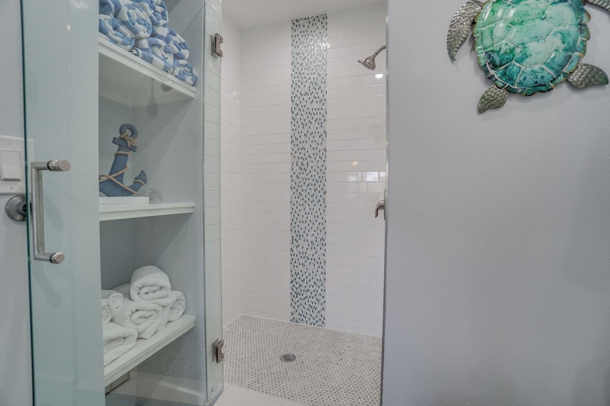 A glass-enclosed shower with a tiled wall, a towel rack with folded towels, and a decorative turtle shell on the wall.