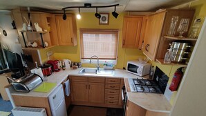 A compact kitchen with wooden cabinets, a sink, a microwave, a stove, and a dining area with a table set for two. - 2-bed Caravan at Billing Aquadrome Northampton (Northampton)