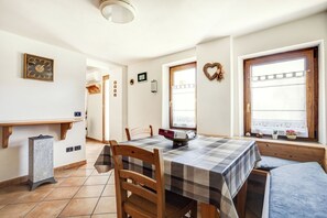 A dining area with a checkered tablecloth, wooden chairs, a wall clock, and a window with a view.